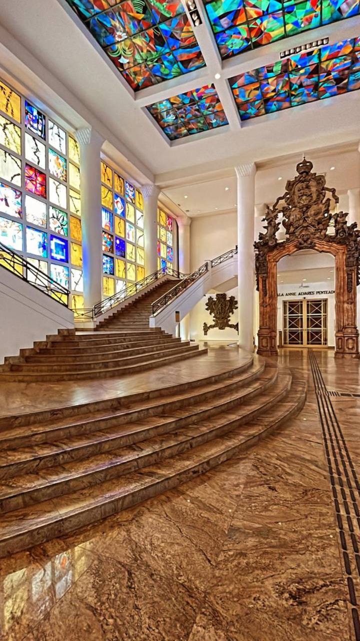 Interior de museu com vitrais coloridos no teto, escada de madeira e porta ornamentada
